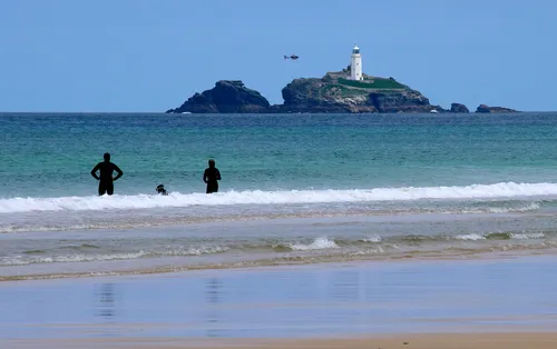 This landscape photograph captures the Godrevy helicopter flying near the iconic Godrevy Lighthouse, which is located off the coast of Cornwall, during a late morning in the summer season. The image shows the turquoise sea and the sandy beach in the foreground, where several people can be seen wading and swimming in the gentle surf. The lighthouse stands prominently atop Godrevy Island, providing a well-known landmark that is often associated with the coastline of Cornwall. The calm sea and bright beach under clear skies emphasize the coastal character of the region, highlighting the connection between the beach, the sea, and the historic Godrevy Lighthouse.